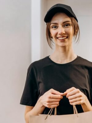 Smiling young woman in black attire holding delivery paper bags indoors.